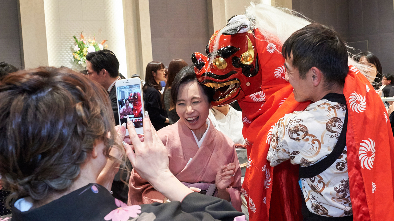 パーティで獅子舞練り歩き。スマホを構える女性とポーズする女性。噛みに行くでん舎の獅子舞。